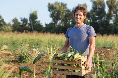 Portrait of smiling farmer holding crate with corn crops on agricultural field