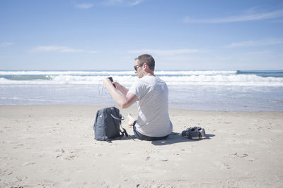 Rear view of man with mobile phone while taking earphones out of backpack at beach