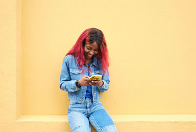 Cheerful african american female teenager with dyed hair wearing denim clothes messaging on mobile phone while sitting near yellow wall