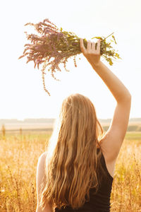 Girl with blond hair stands with her back, raises her hands up and holds a bouquet of wildflowers