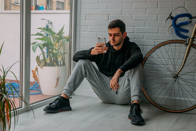 Young man using mobile phone while sitting against wall outdoors