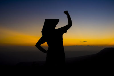 Silhouette young woman gesturing while standing on mountain against sky during sunset