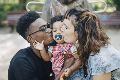 Mother and father kissing daughter sucking pacifier at playground