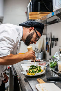 Side view of busy male chef in uniform adding sauce on top of dish with greenery while working in kitchen of cafe