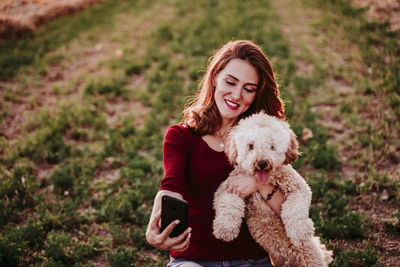 Woman carrying dog while taking selfie on grassy field