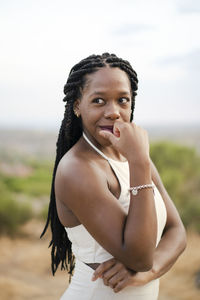 Adult black female in white top with afro braids touching face and looking away on blurred background of countryside