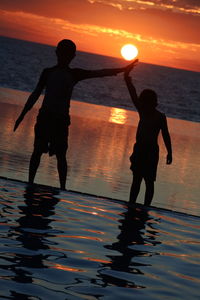 Silhouette boys standing by infinity pool against sea during sunset