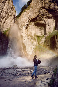 Woman in jeans and boots stands next to a waterfall in the summer in the caucasus