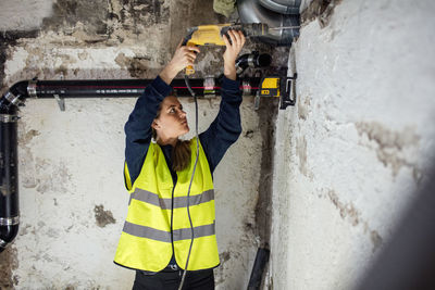 Confident woman drilling on wall above pipes at basement