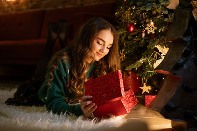 Happy teenage girl opens red gift box sitting under christmas tree at midnight