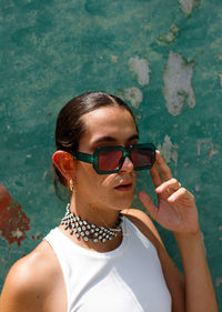 Confident young ethnic female model with dark hair touching fashionable sunglasses and looking away against shabby wall on sunny day