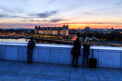 People on illuminated city by river against sky during sunset