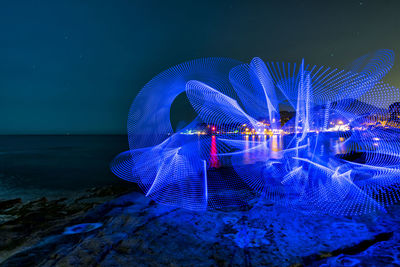 Illuminated ferris wheel by sea against sky at night