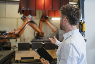 Mature businessman with digital tablet gesturing toward robotic arm machine while standing at factory