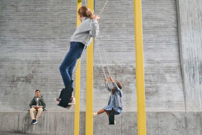 Carefree sisters swinging on swings at playground while brother sitting in background