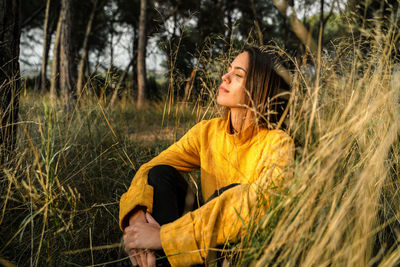 Tranquil female sitting in dry field and enjoying sunset in countryside with closed eyes while embracing knees