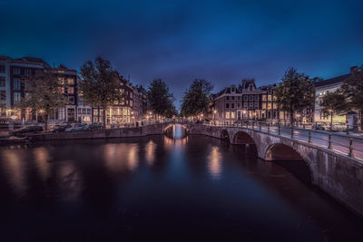 Bridge over river by illuminated buildings against sky in city