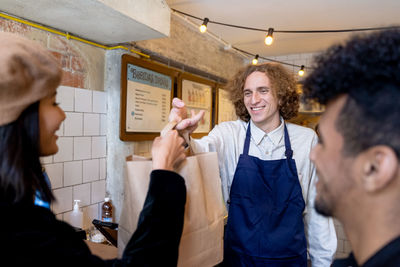 Smiling young male server in apron passing takeaway food to ethnic couple while standing at restaurant counter