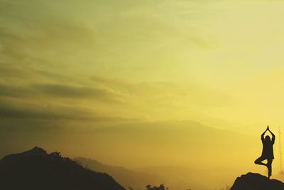 Silhouette man in tree pose on cliff against orange sky