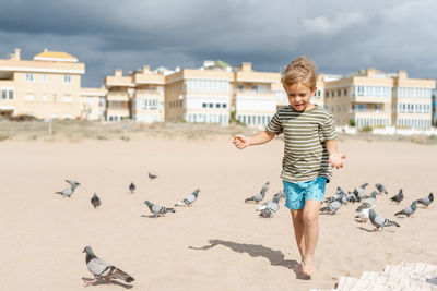 A boy feeds pigeons on a sandy beach against a background of apartment complexes and a stormy sky