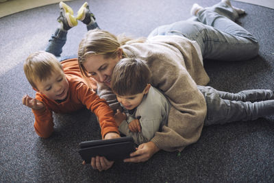 High angle view of kids watching digital tablet with female teacher lying down on carpet at preschool