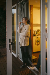 Young woman wearing sweater brushing teeth while standing at doorway seen through glass door