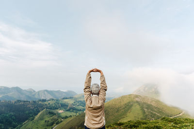 Back view of unrecognizable senior female traveler raising and stretching arms in morning in hilly terrain