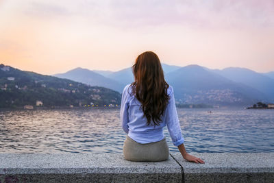 Rear view of woman sitting by sea against mountains during sunset