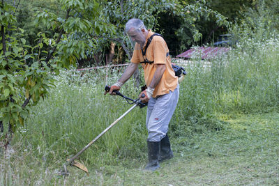 A mature man in protective clothing, and gloves  and mowing tall grass and the weeds with a trimmer