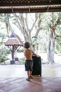 Rear view of shirtless boy pushing wheeled luggage on porch at resort