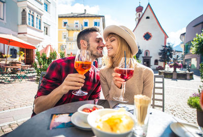 Smiling couple holding wineglass embracing while sitting at cafe outdoors