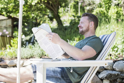 Side view of smiling man reading newspaper on lounge chair at yard