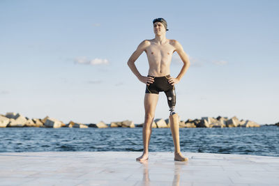 Young man with hands on hip looking away while standing against sea during sunny day