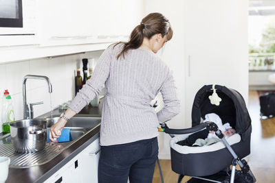 Rear view of woman looking at baby girl in carriage while washing utensils in kitchen