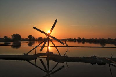 Silhouette windmill by lake against sky during sunset