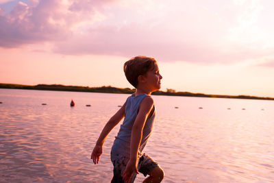 Side view of boy running at beach against sky during sunset