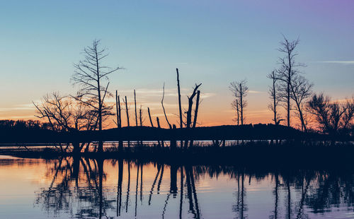 Silhouette bare trees by lake against sky during sunset
