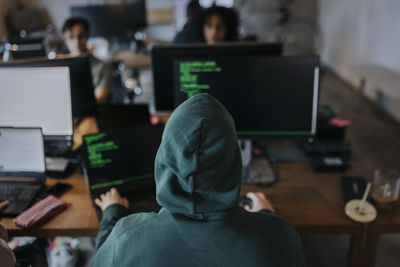 Rear view of young male hacker wearing hood coding on computer at desk in creative workplace