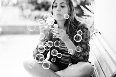 Young woman blowing soap bubbles while sitting on bench