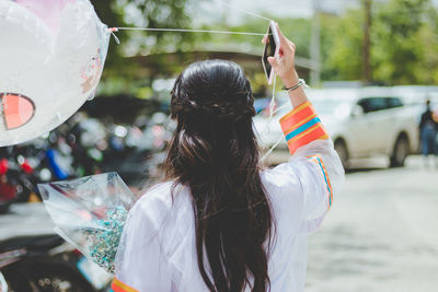 Rear view of woman holding balloon and mobile phone while standing in city