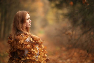 Thoughtful young female model covered with autumn leaves in forest