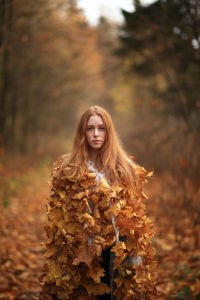 Portrait of young woman covered with leaves standing at forest during autumn