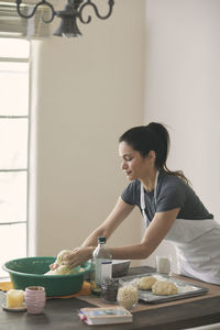 Woman in apron kneading dough on table by wall at home