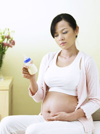 Pregnant woman looking at milk bottle while sitting on sofa at home