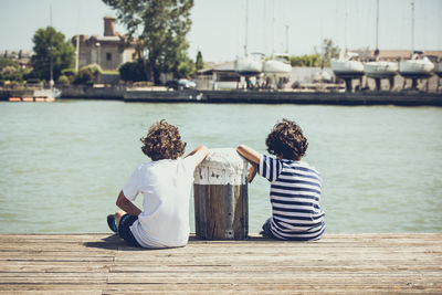 Rear view of boys sitting on pier against river during sunny day