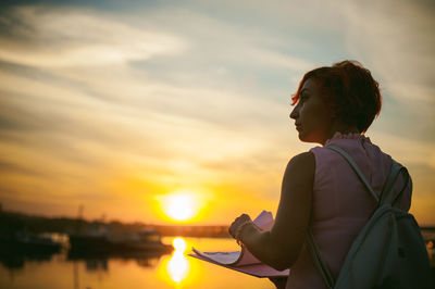 Rear view of mid adult woman holding papers while standing by river against sky during sunset