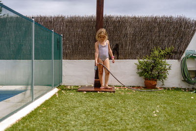 Cute little girl in swimsuit walking and watering green lawn from hose during summer holidays in countryside