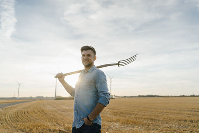 Portrait of smiling young man holding pitchfork standing on stubble field