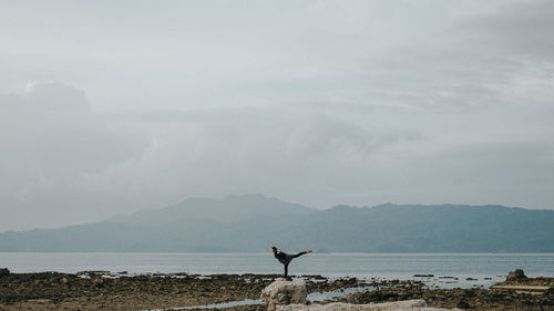 Man doing yoga on rock by sea against sky