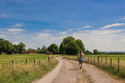 Rear view of woman walking on track through field towards trees with a blue sky.